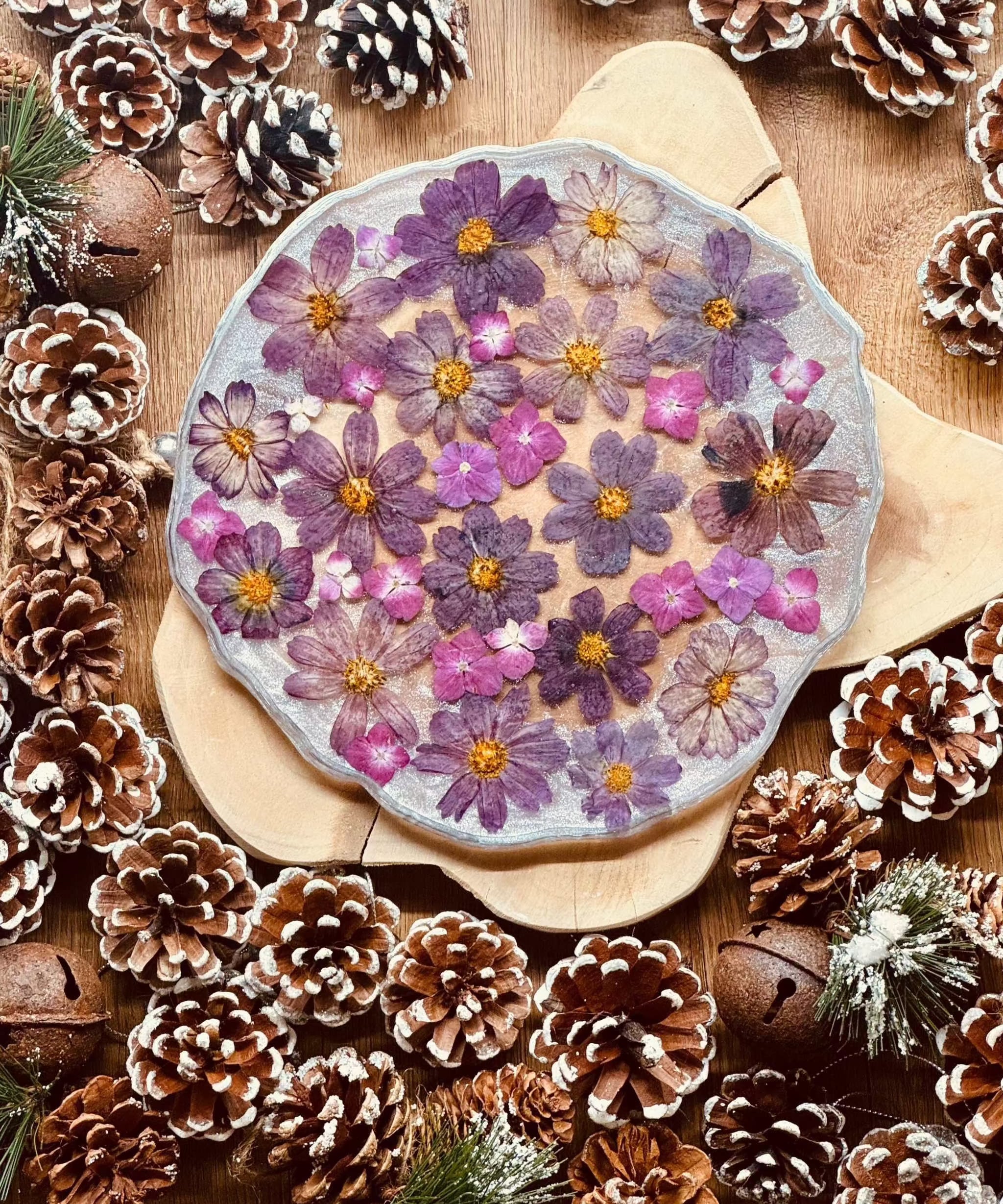 Handgemachtes Tablett aus Resin mit eingebetteten lila Blüten und silbernen Details auf Holzunterlage umgeben von Tannenzapfen und Weihnachtsdekoration