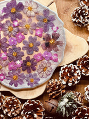 Handgemachtes Tablett aus Resin mit eingebetteten lila Hortensienblüten und silbernen Details, dekorativ auf Holzunterlage mit Kiefernzapfen arrangiert