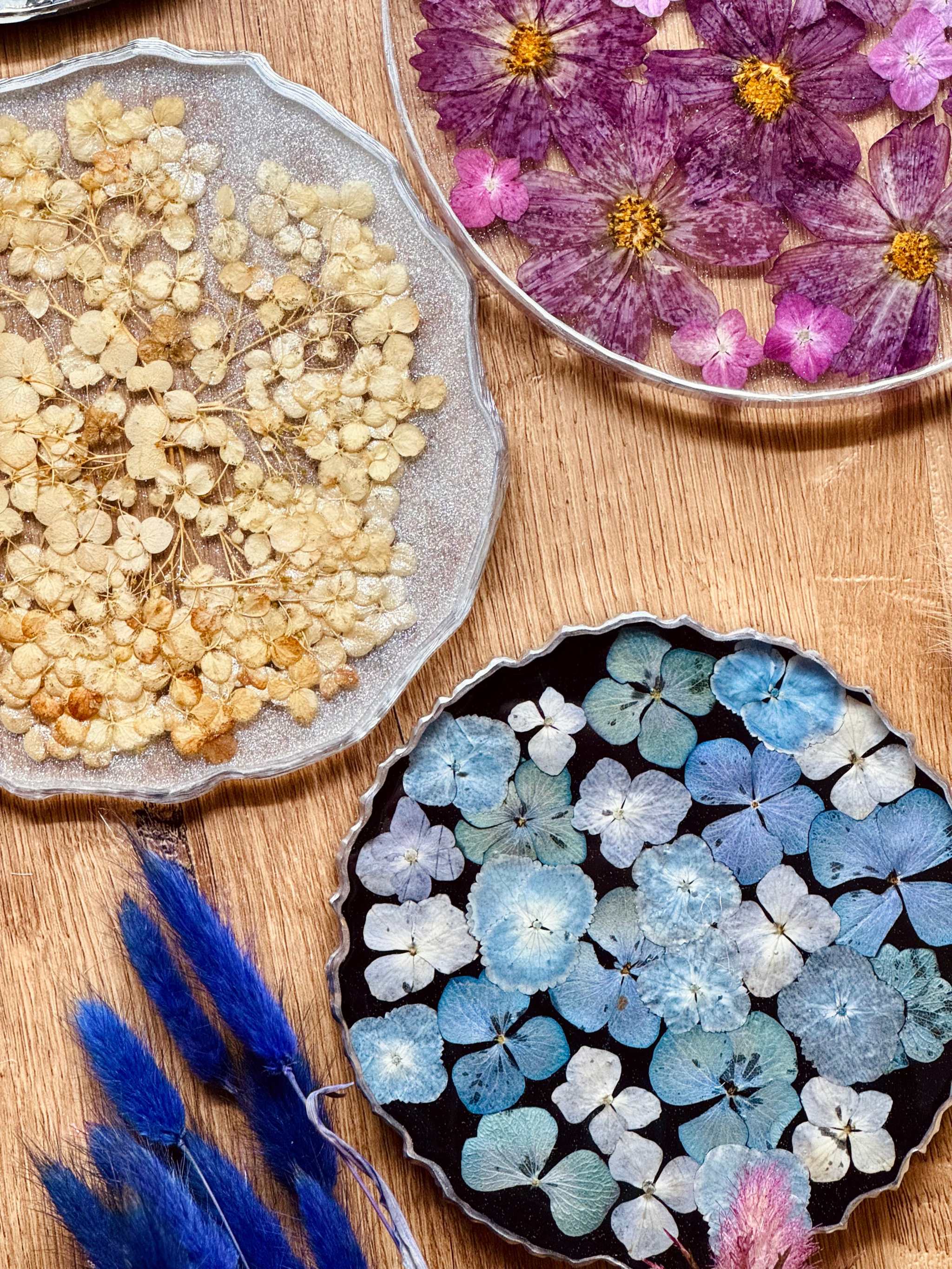 handgemachtes Tablett aus Resin mit getrockneten Blumen in Blau, Lila und Beige sowie silbernen Details auf Holzuntergrund