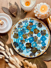 rundes handgemachtes Tablett aus Resin mit blauen Blumen und goldenen Details auf Holztisch mit Kerzen und Dekoration