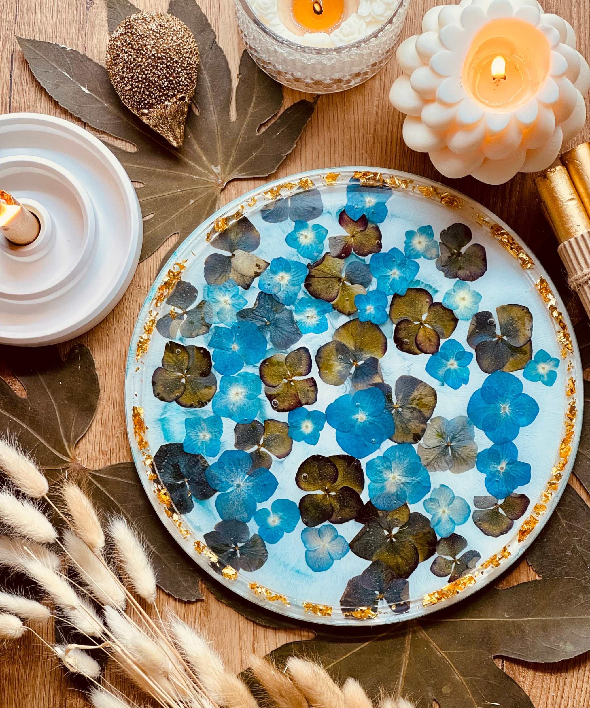 rundes handgemachtes Tablett aus Resin mit blauen Blumen und goldenen Details auf Holztisch mit Kerzen und Dekoration