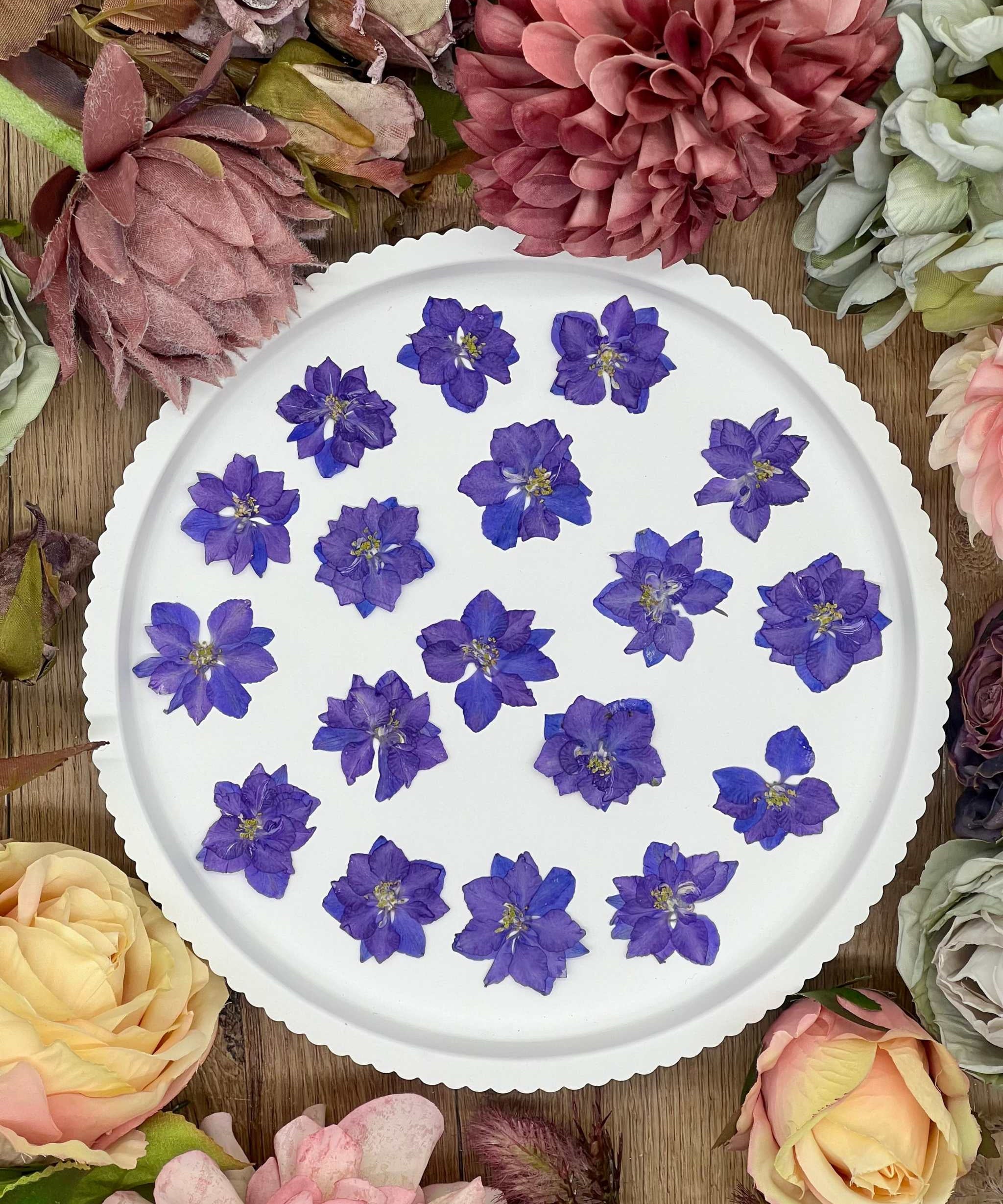 12 purple dried Larkspur flowers on a white plate surrounded by various blooms.