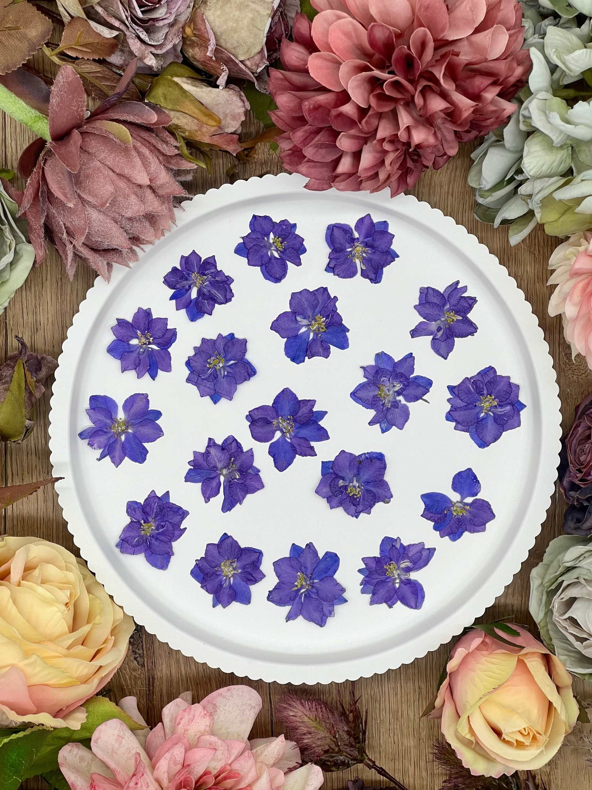 12 purple dried Larkspur flowers on a white plate surrounded by various blooms.