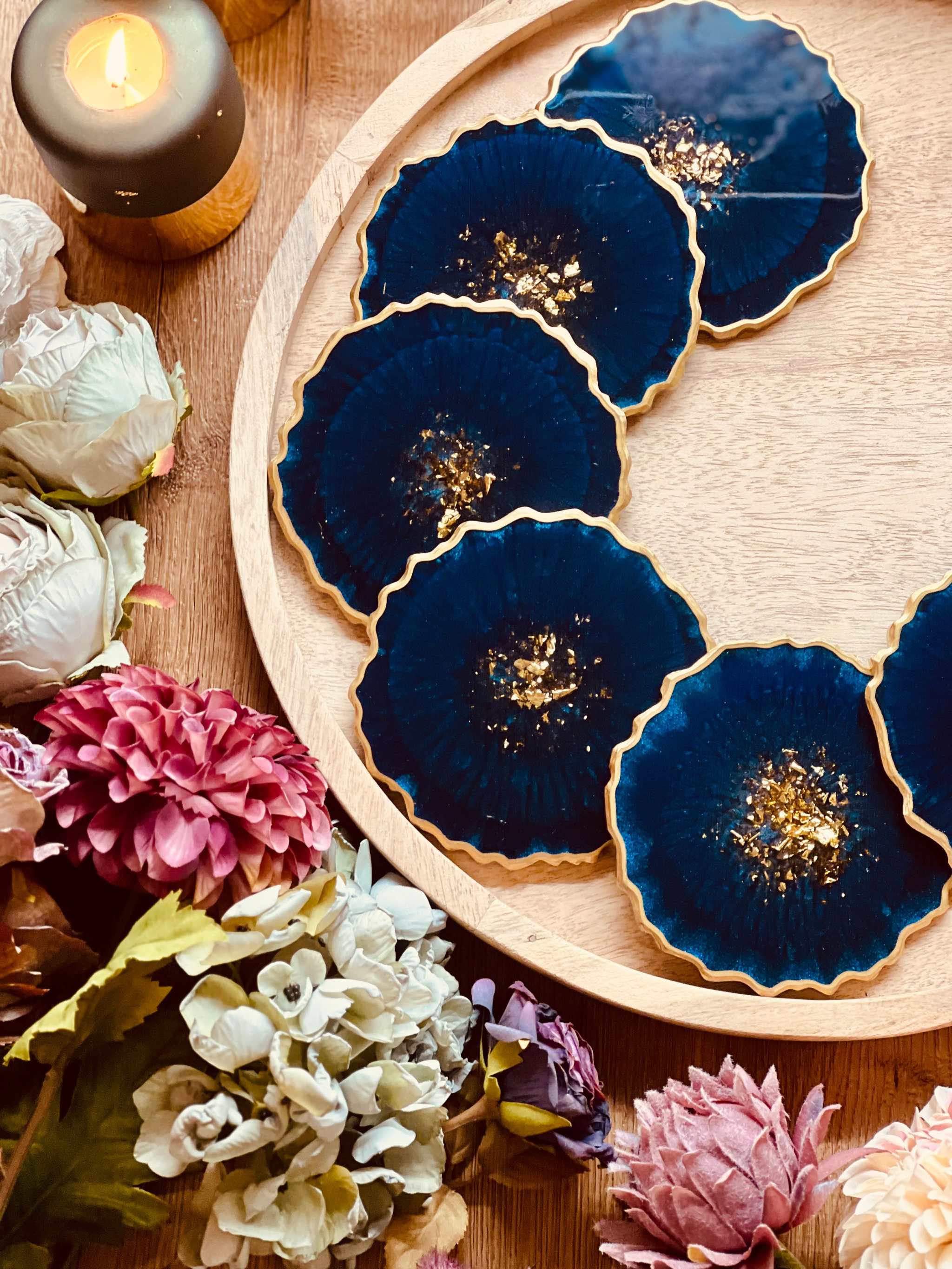 6 handgemachte blaue Resin Untersetzer mit goldenen Details auf Holztablett, umgeben von Blumen und Kerze