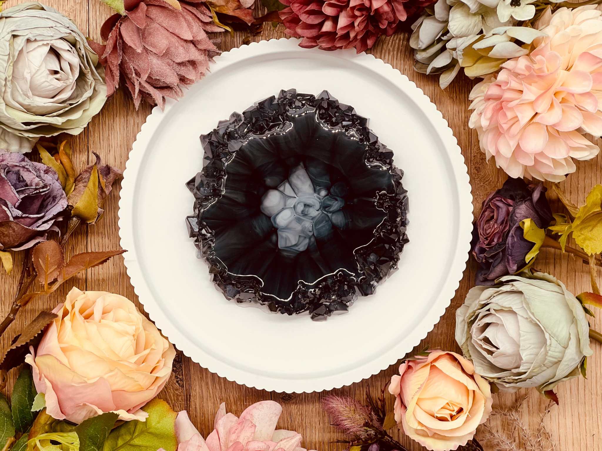 Rundes handgemachtes Tablett aus Resin im schwarzen Geode Kristall Design umgeben von künstlichen Blumen auf Holzuntergrund