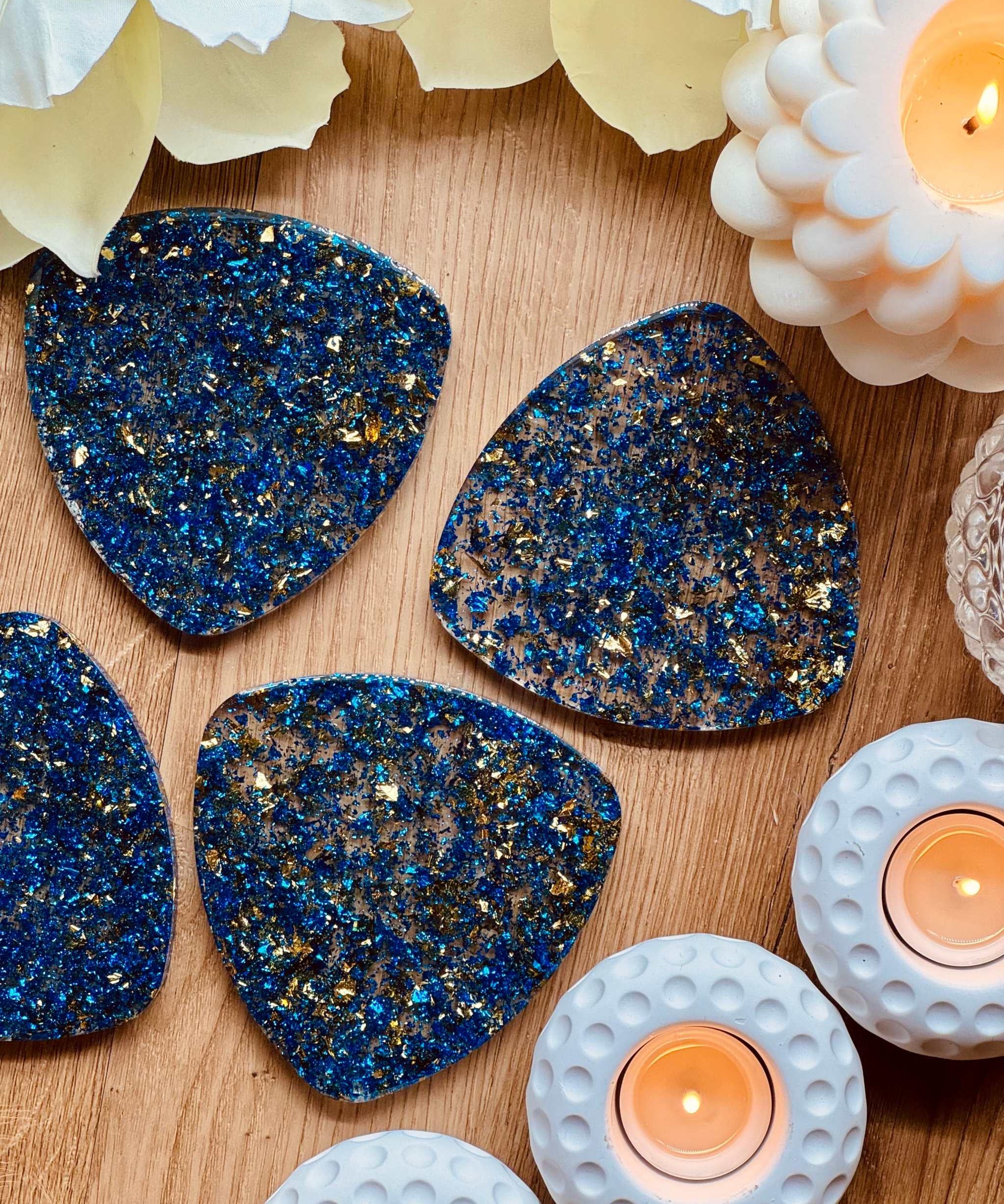 4 Untersetzer aus blauem Resin mit goldenen Details liegen dekorativ auf Holztisch neben Kerzen und Blüten
