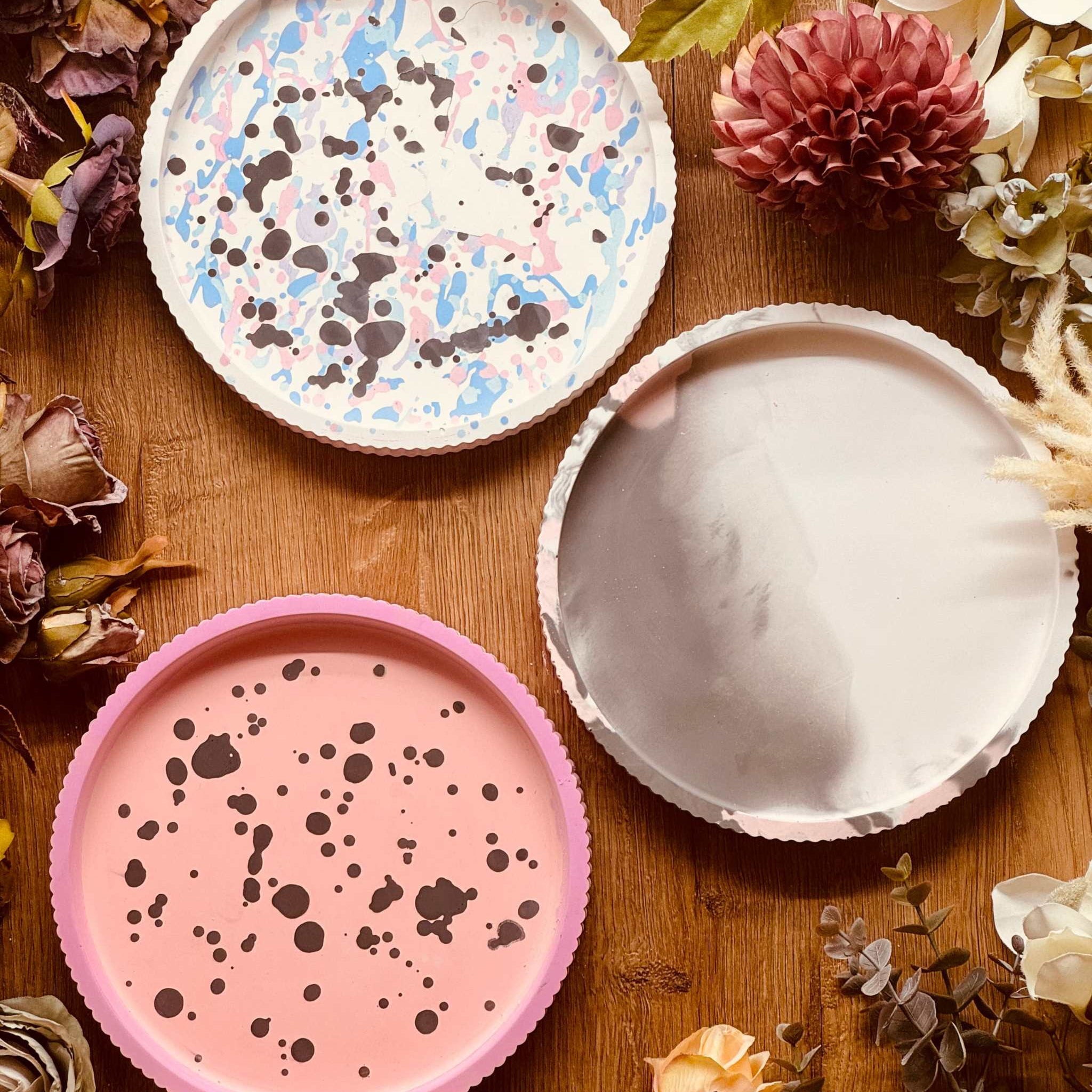 Drei handgefertigte Gießkeramik Tabletts mit einzigartigen Mustern: rosa mit schwarzen Sprenkeln, pastellfarben gesprenkelt, und in dezentem Marmor-Grau. Perfekt als Dekoration oder Serviertablett.Giesskeramik Weisse Deko Tablett buntes Design modern