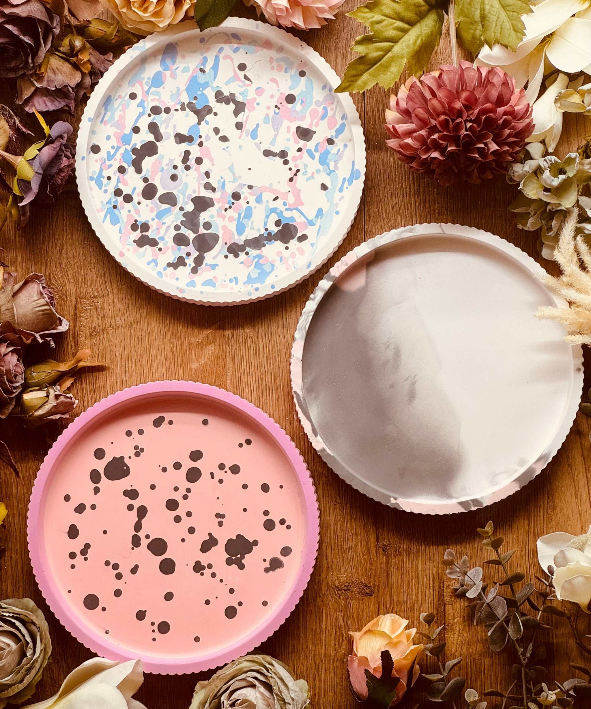 Drei handgefertigte Gießkeramik Tabletts mit einzigartigen Mustern: rosa mit schwarzen Sprenkeln, pastellfarben gesprenkelt, und in dezentem Marmor-Grau. Perfekt als Dekoration oder Serviertablett.Giesskeramik Weisse Deko Tablett buntes Design modern