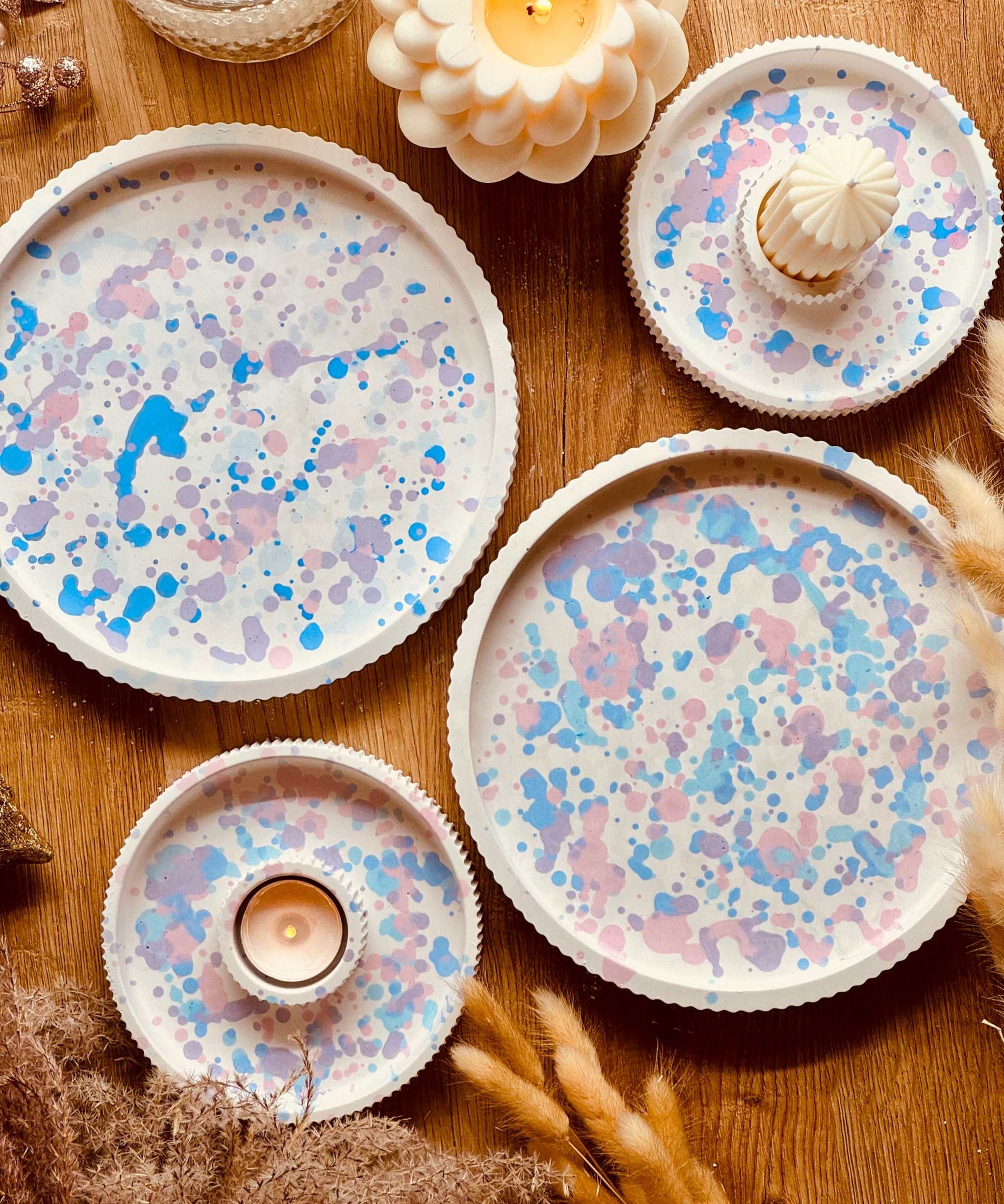 Set aus Gießkeramik-Tabletts in verschiedenen Größen mit pastellfarbenem Sprenkel-Design in Rosa, Blau und Flieder, kombiniert mit handgefertigten Kerzenhaltern, ideal für eine stilvolle Tischdekoration.
