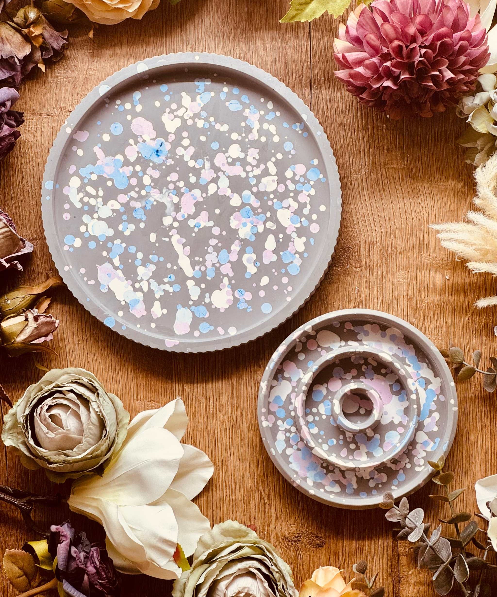 Großes rundes Tablett und passender Stabkerzenhalter aus Gießkeramik im modernen Sprenkel-Design, in sanften Pastelltönen wie Blau, Rosa und Weiß auf grauem Hintergrund, umgeben von Trockenblumen.