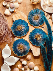4 handgemachte blaue Resin-Untersetzer mit goldenen Details auf Holztisch und Muscheldeko