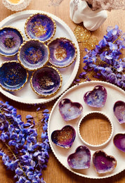 Handgemachte lila Schmuckschalen aus Resin mit goldenen Details in runder und herzförmiger Form auf Holzuntergrund mit lila Blumenarrangement
