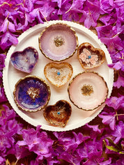 Schmuckschalen aus Resin in Beerentönen mit goldenen Details, handgemachte Unikate auf weißem Teller, umgeben von violetten Blüten.