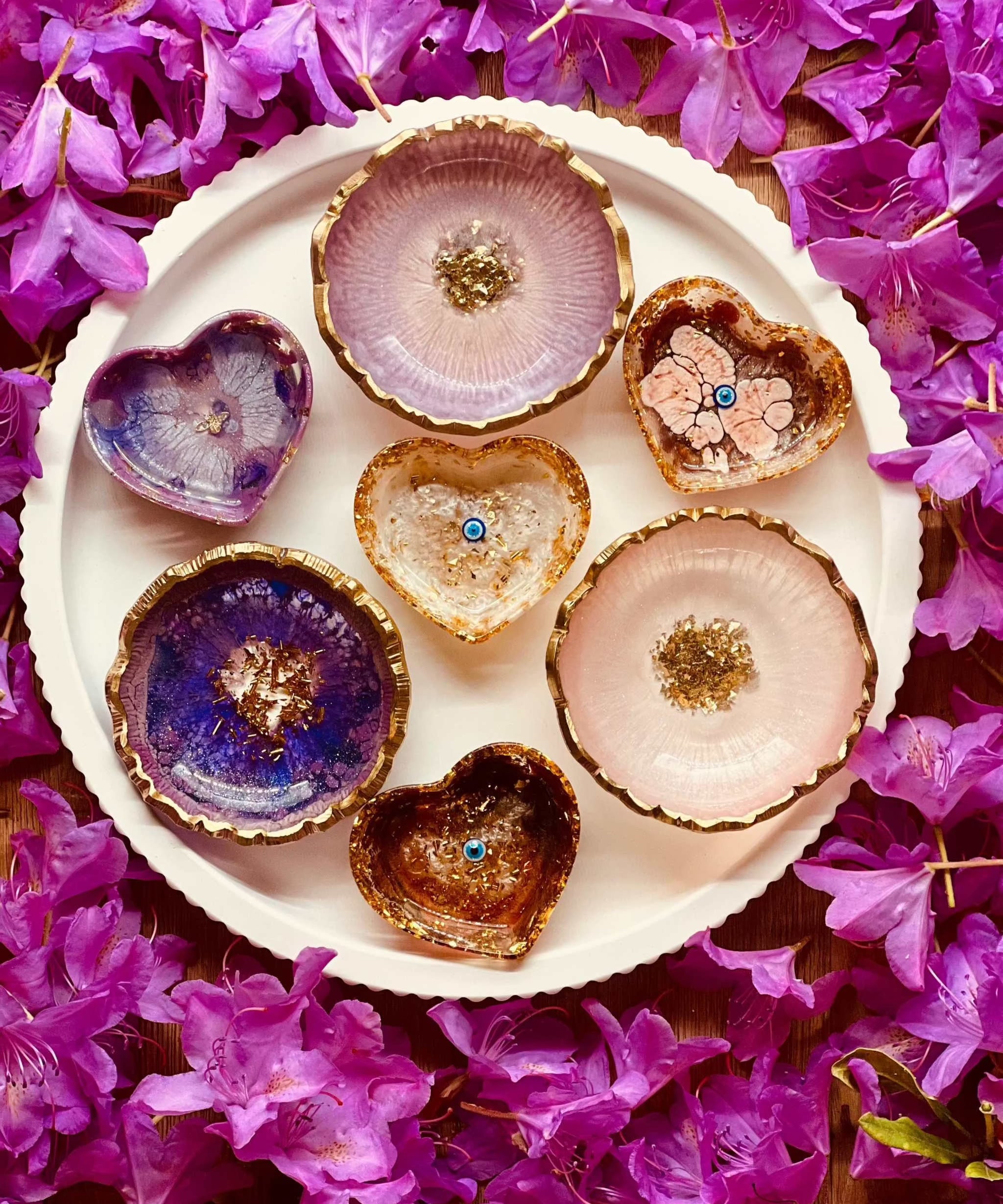 Schmuckschalen aus Resin in Beerentönen mit goldenen Details, handgemachte Unikate auf weißem Teller, umgeben von violetten Blüten.