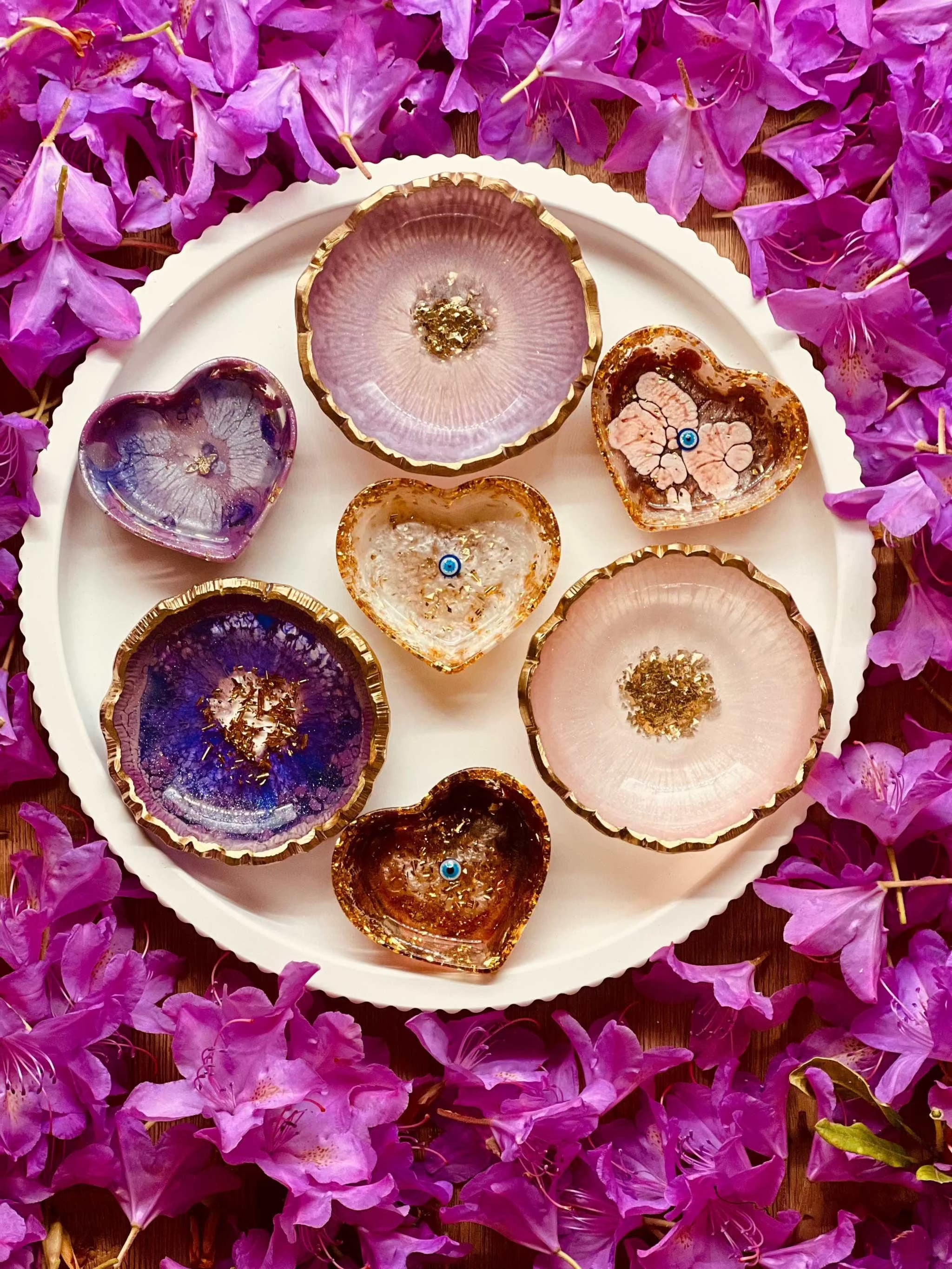 Schmuckschalen aus Resin in Beerentönen mit goldenen Details, handgemachte Unikate auf weißem Teller, umgeben von violetten Blüten.