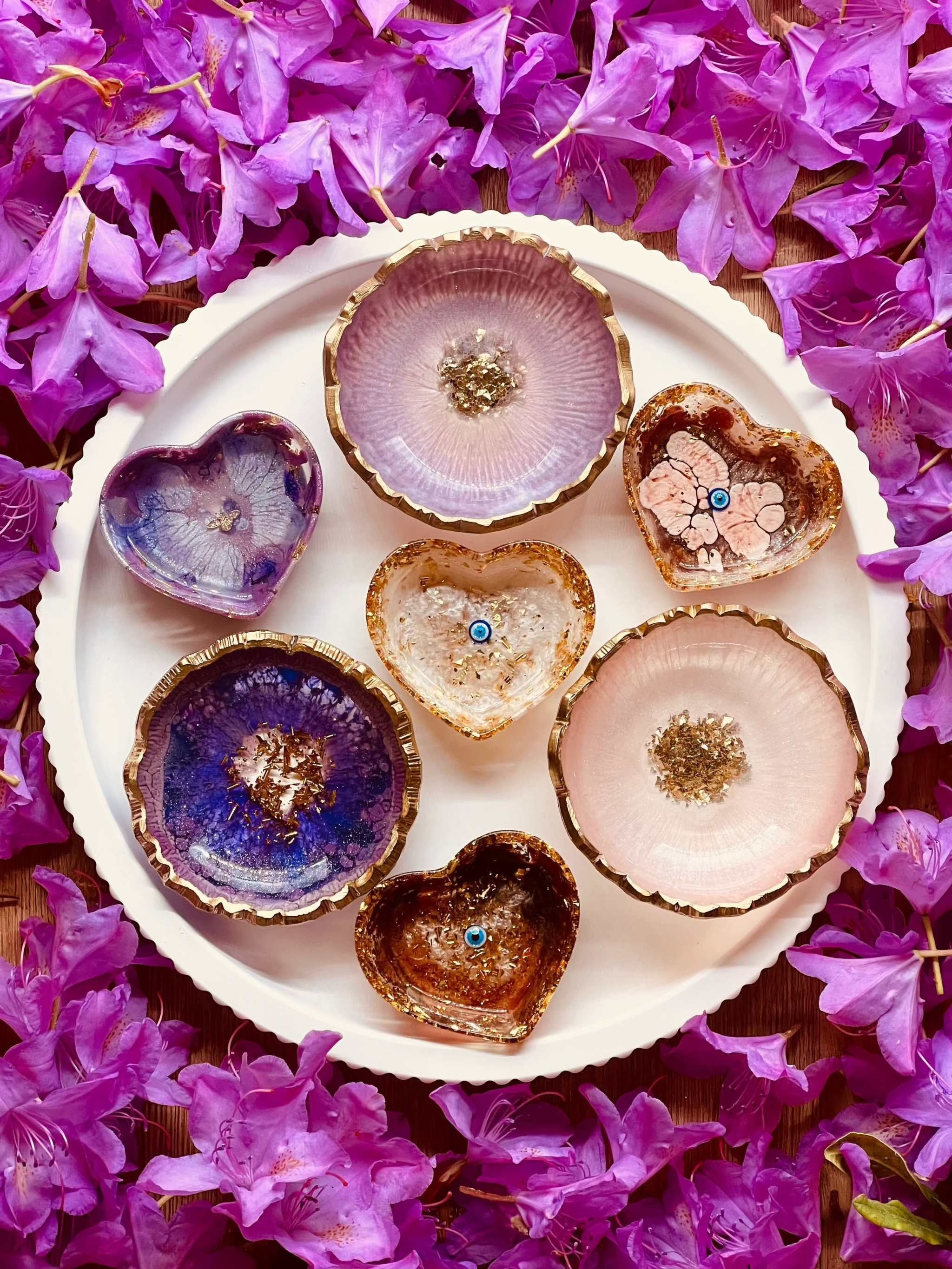 Schmuckschalen aus Resin in Beerentönen mit goldenen Details auf weißem Teller, umgeben von pinken Blüten.