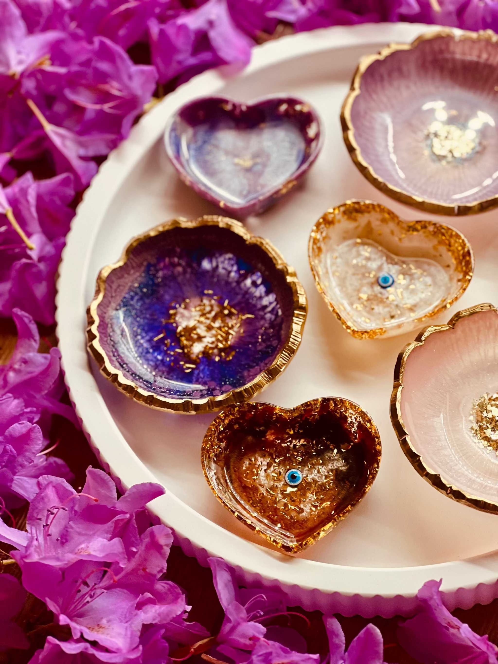Handgemachte Schmuckschalen aus Resin in Beerentönen mit goldenen Details auf weißem Tablett neben lila Blüten