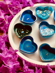 Herzförmige handgemachte Schmuckschalen aus Resin in verschiedenen Blau- und Grüntönen mit silbernen Details auf weißem Tablett zwischen pinken Blüten