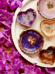Handgemachte Schmuckschalen aus Resin in Beerentönen mit goldenen Details und unterschiedlichen Formen auf weißem Teller mit pinken Blüten