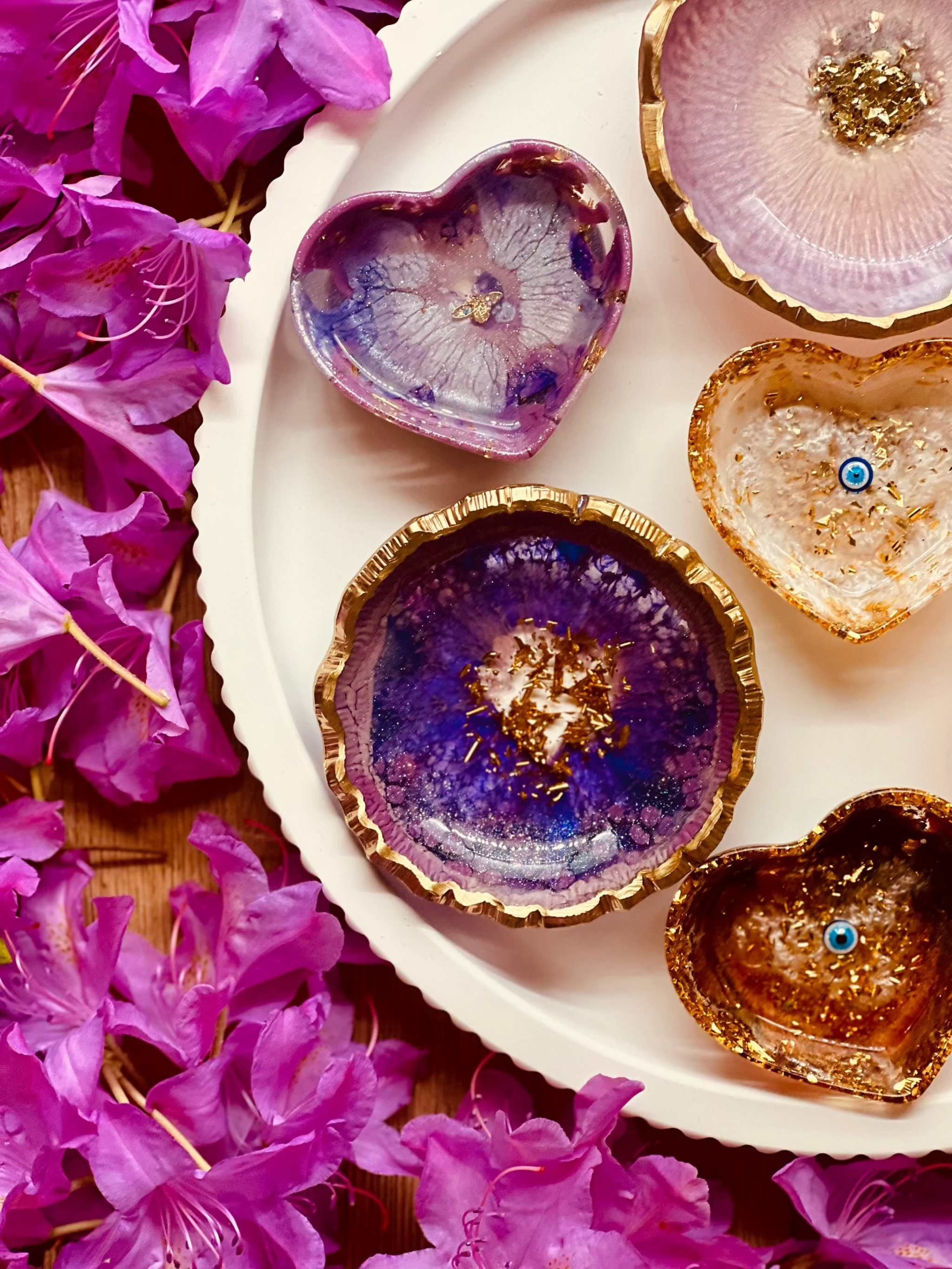 Handgemachte Schmuckschalen aus Resin in Beerentönen mit goldenen Details und unterschiedlichen Formen auf weißem Teller mit pinken Blüten