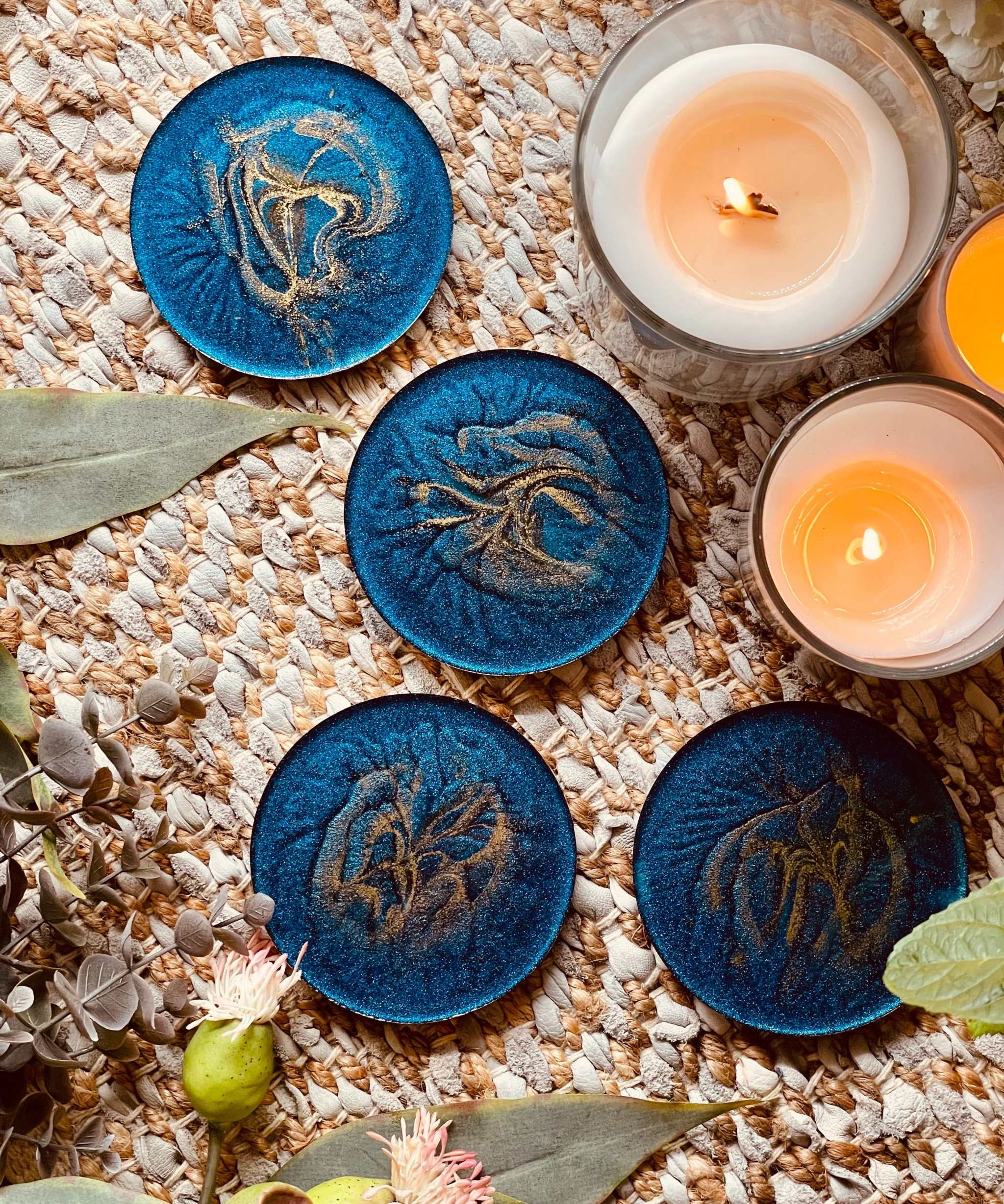 4 handgemachte Untersetzer aus blauem Resin mit goldenen Details auf Teppich neben Kerzen und Blumen