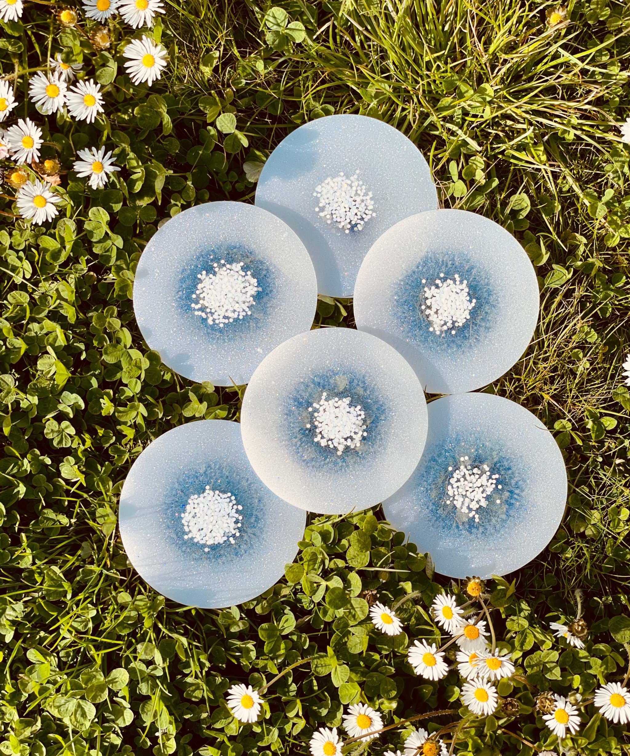 Sechs handgemachte weiße Untersetzer aus Resin mit glitzernden Details auf Wiese mit Blumen