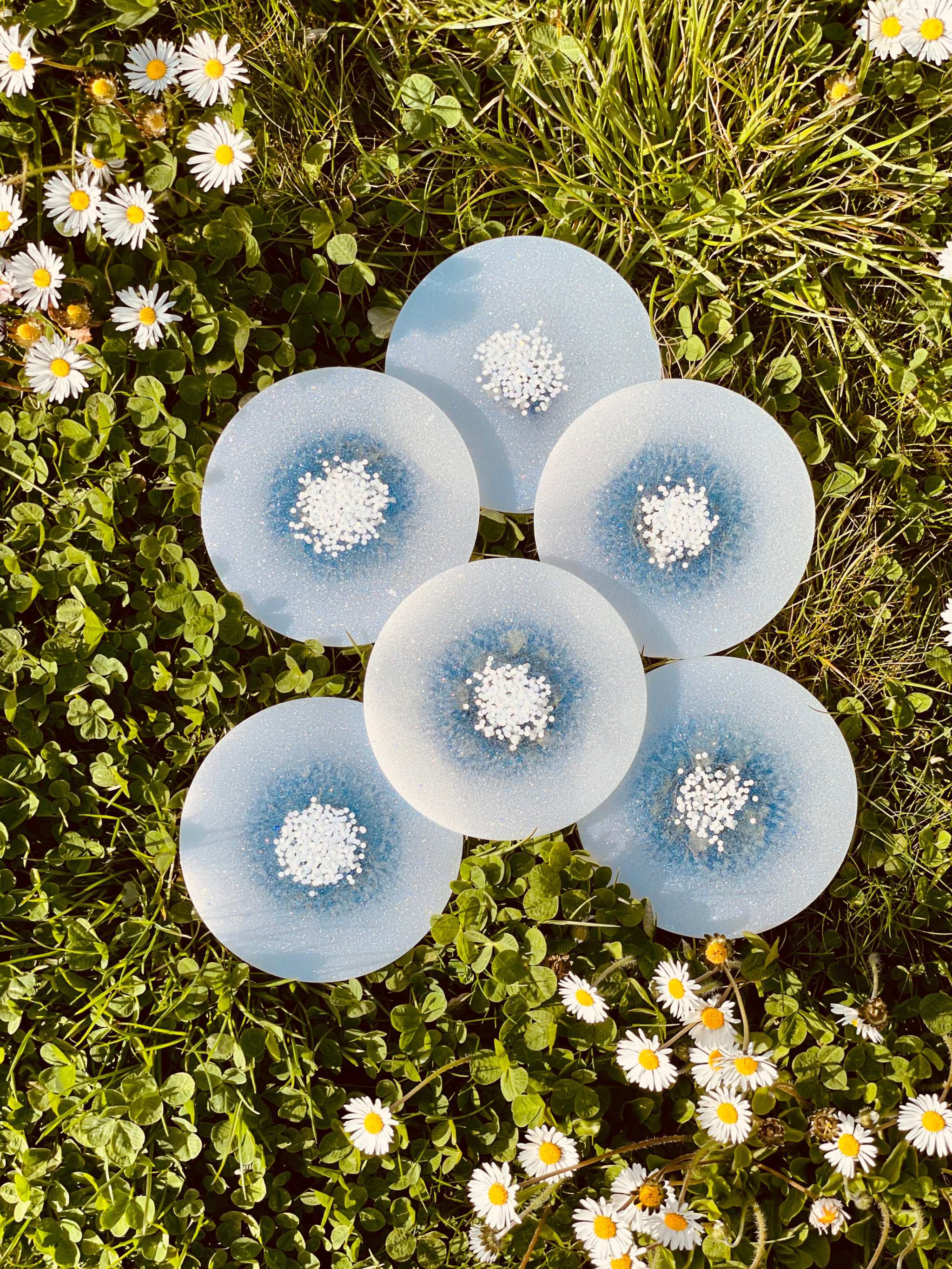 Sechs handgemachte weiße Untersetzer aus Resin mit glitzernden Details auf Wiese mit Blumen