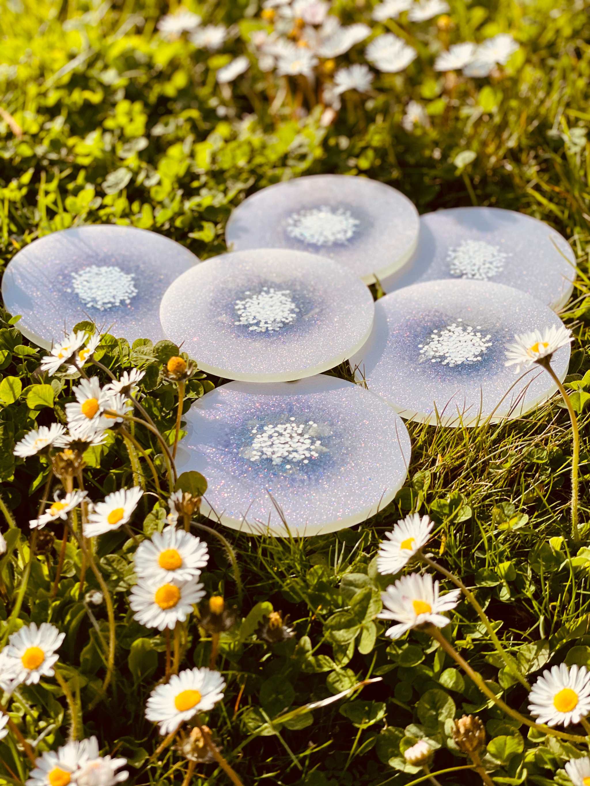 6 handgemachte weiße Untersetzer aus Resin mit glitzernden Details auf Wiese mit Blumen
