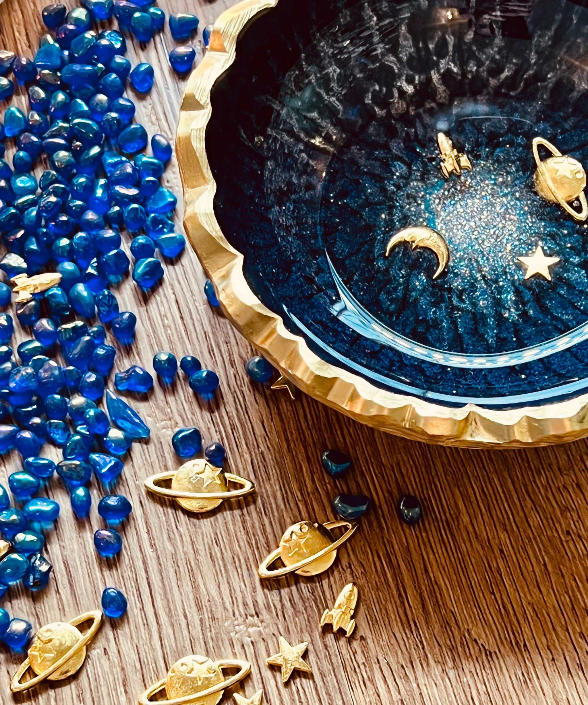 Dunkelblaue handgemachte Schmuckschale aus Resin mit goldenen Galaxy-Details und gezacktem Rand, umgeben von blauen Perlen und goldenen Weltraumfiguren.