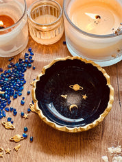 dunkelblaue handgemachte Schmuckschale aus Resin mit goldenen Galaxy Details und gezacktem Rand, dekoriert mit kleinen goldenen Planeten und Sternen, neben Kerzen und blauen Dekosteinen auf Holztisch