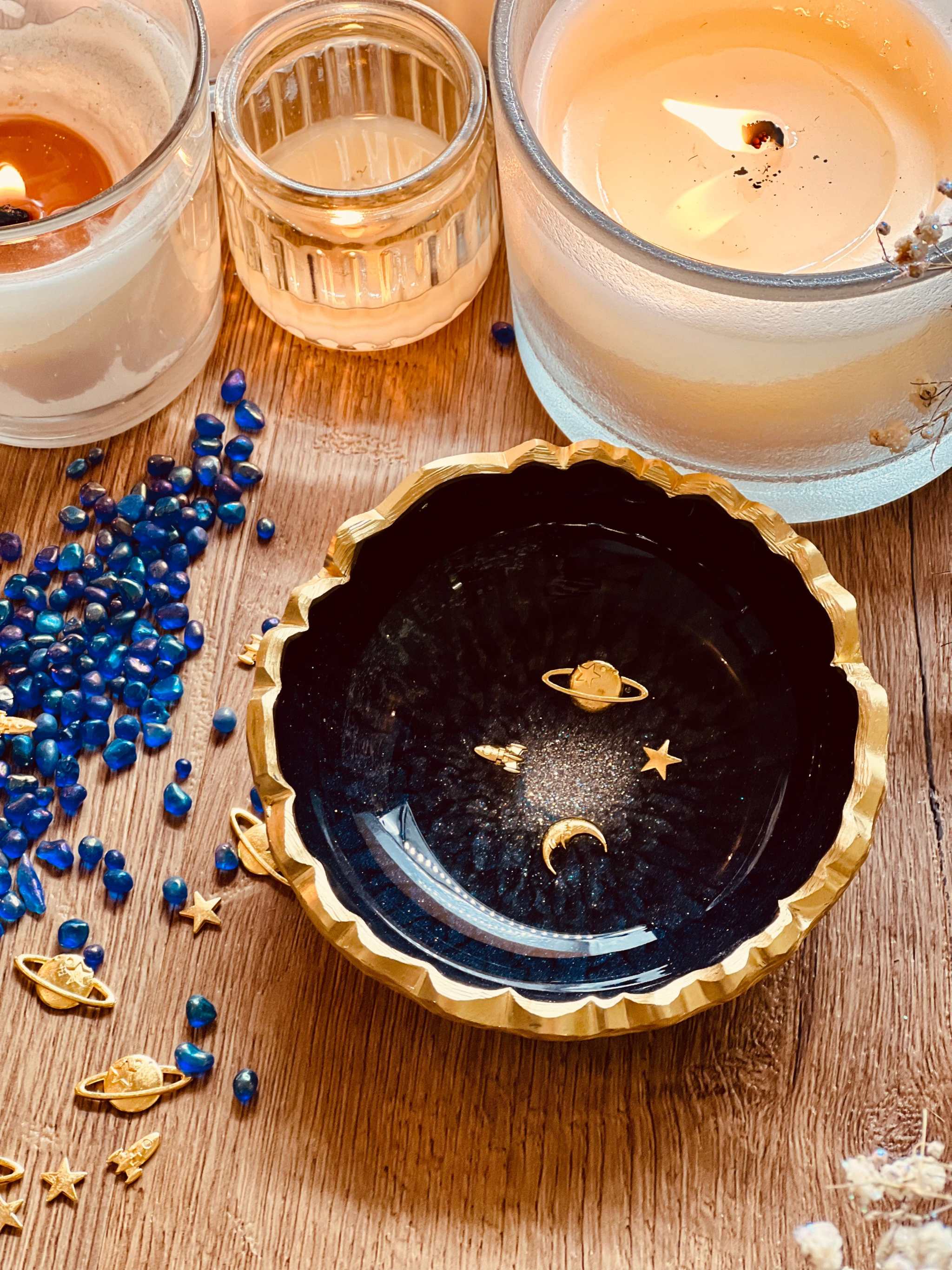 dunkelblaue handgemachte Schmuckschale aus Resin mit goldenen Galaxy Details und gezacktem Rand, dekoriert mit kleinen goldenen Planeten und Sternen, neben Kerzen und blauen Dekosteinen auf Holztisch