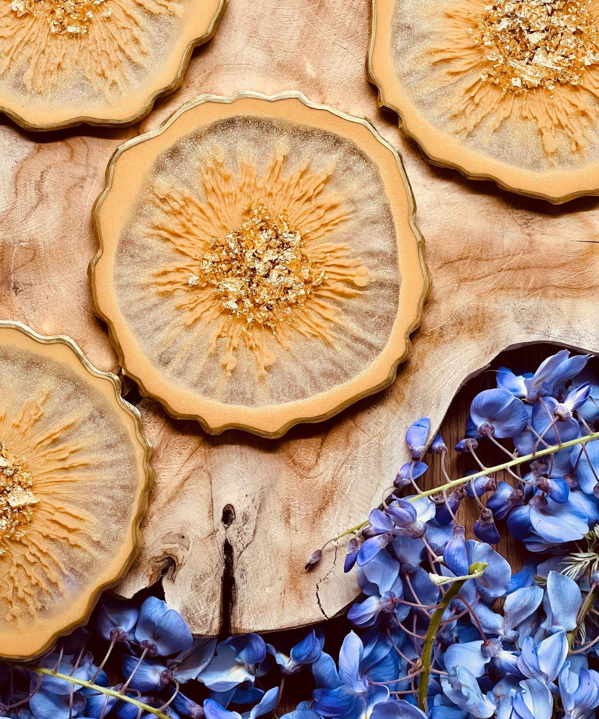 4 handgemachte beige Resin Untersetzer mit goldenen Details auf Holzuntergrund neben blauen Blüten