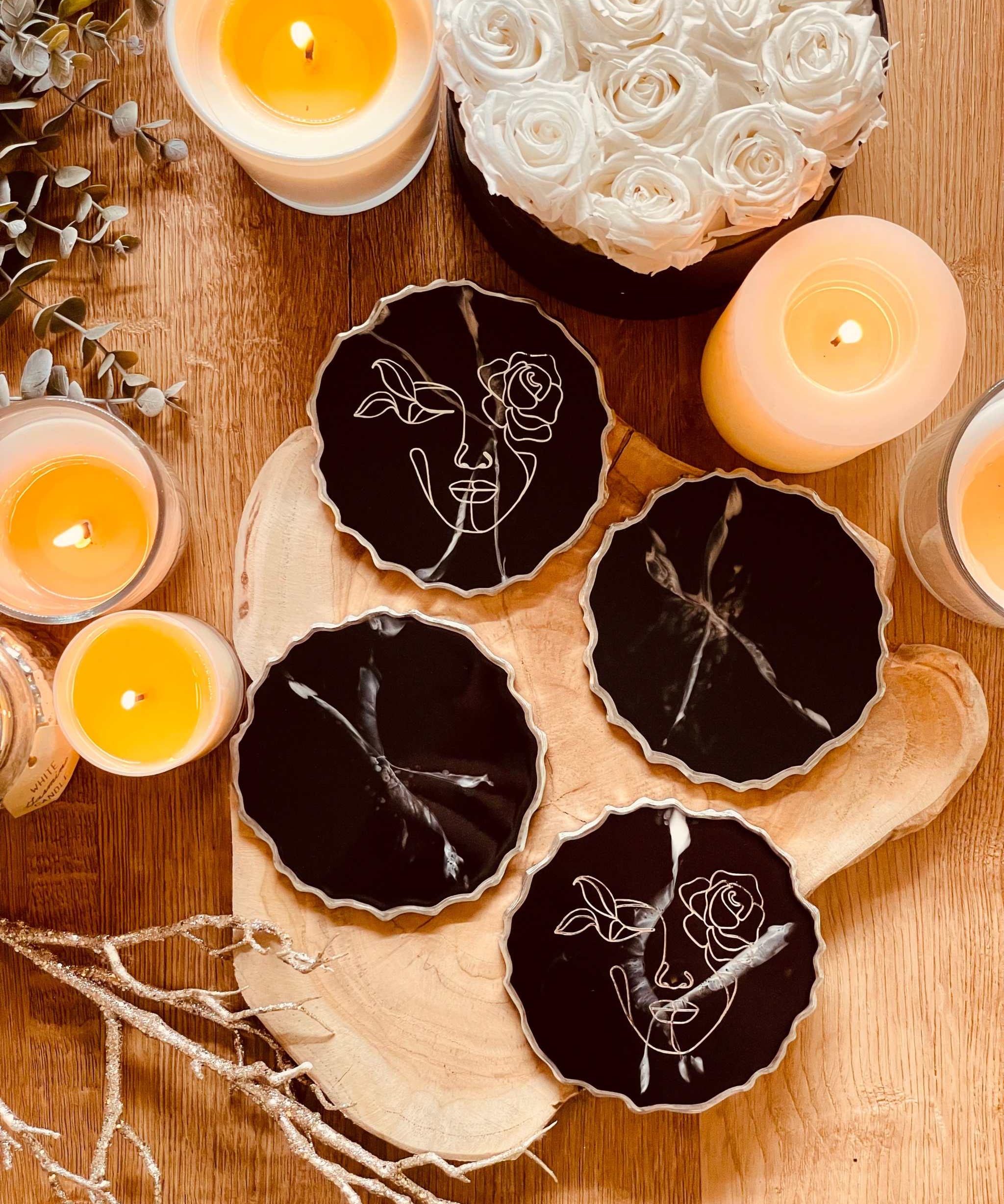 Vier handgemachte Untersetzer aus schwarzem Resin mit silberner LineArt, dekorativ auf Holzunterlage mit Kerzen und weißen Rosen.