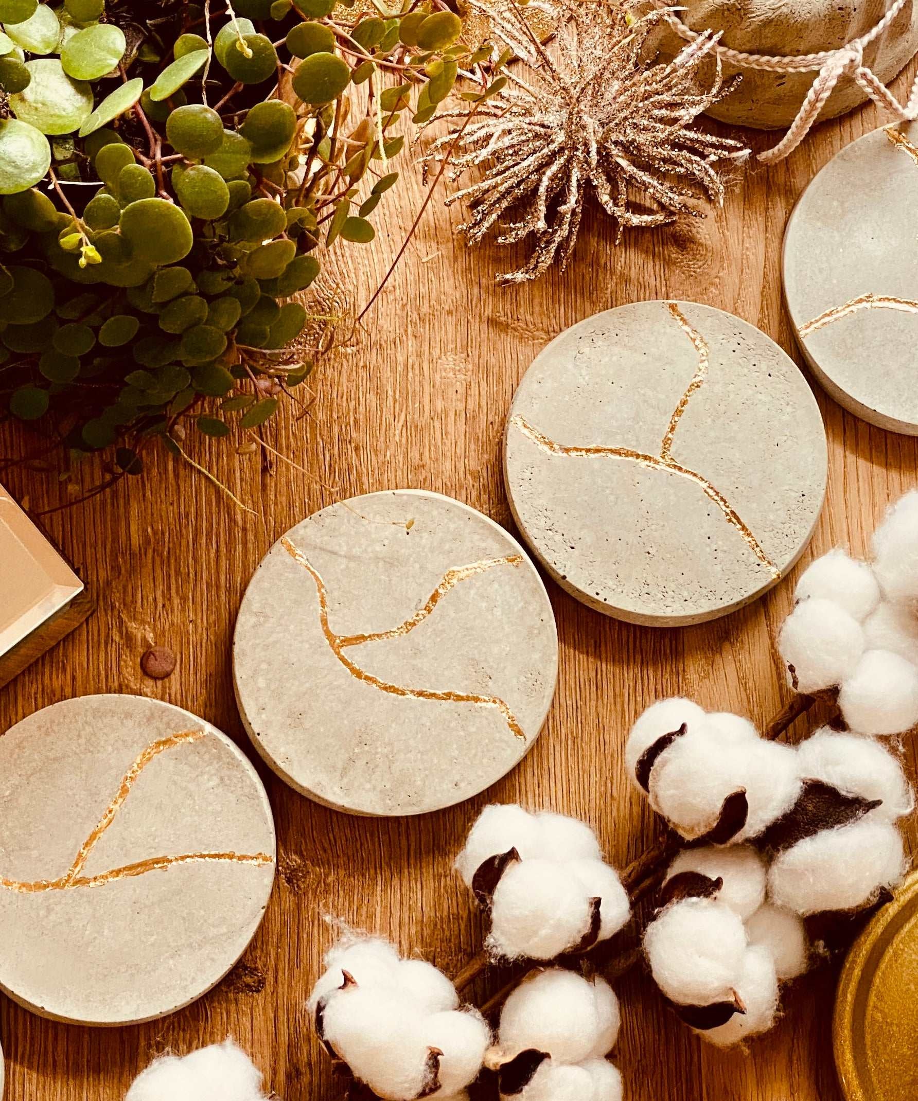 Vier runde Untersetzer aus Beton mit goldener Kintsugi-Verzierung, handgemacht und dekorativ auf Holztisch mit Pflanzen und Baumwollzweigen.
