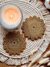 Zwei handgemachte goldene Mandala Untersetzer aus Resin auf einem gewebten Platzset neben einer brennenden Kerze