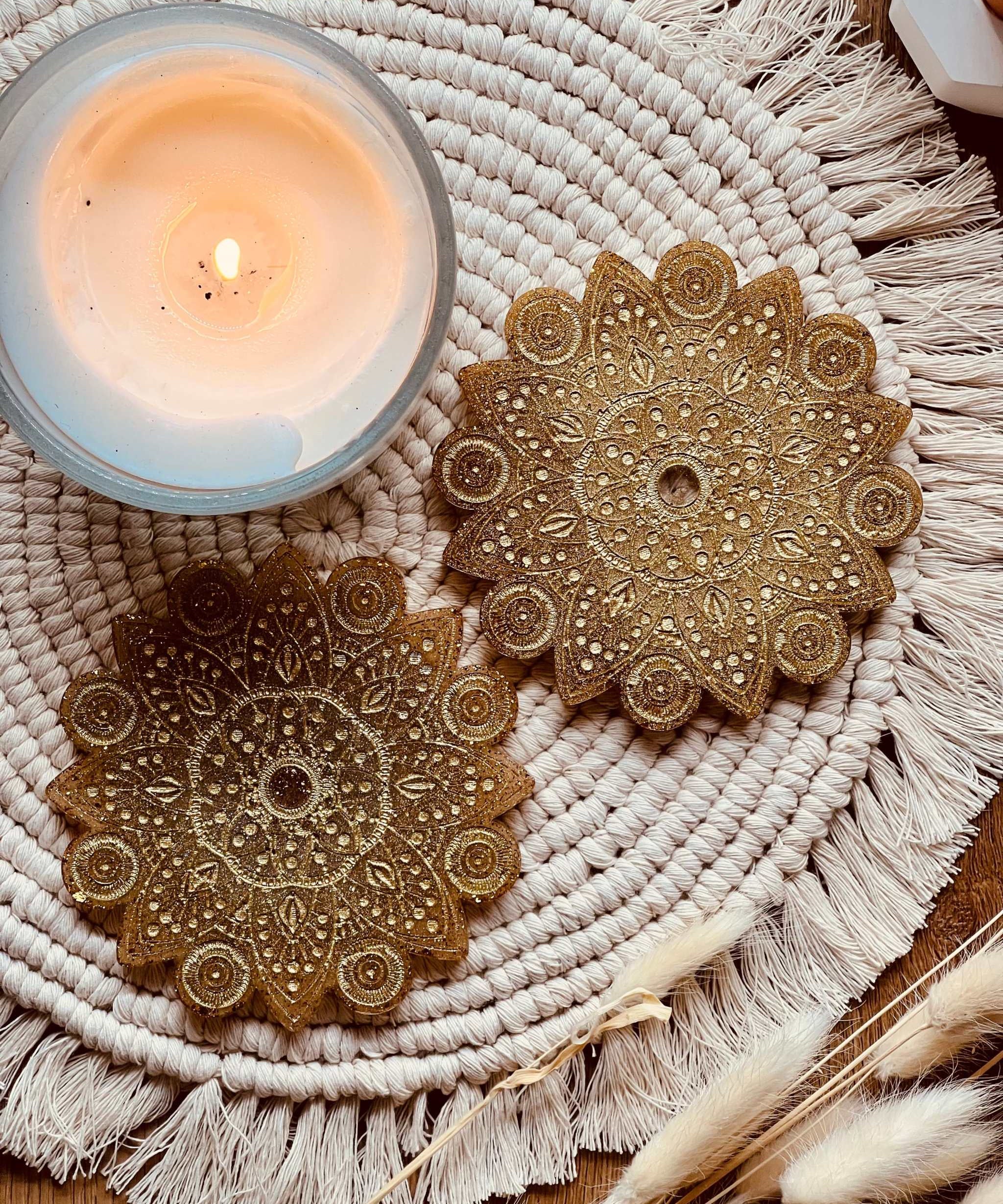 Zwei handgemachte goldene Mandala Untersetzer aus Resin auf einem gewebten Platzset neben einer brennenden Kerze