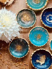 Handgemachte Schmuckschalen aus Resin in Blau, Türkis und Grün mit goldenen Details auf Holztisch neben weißer Blume