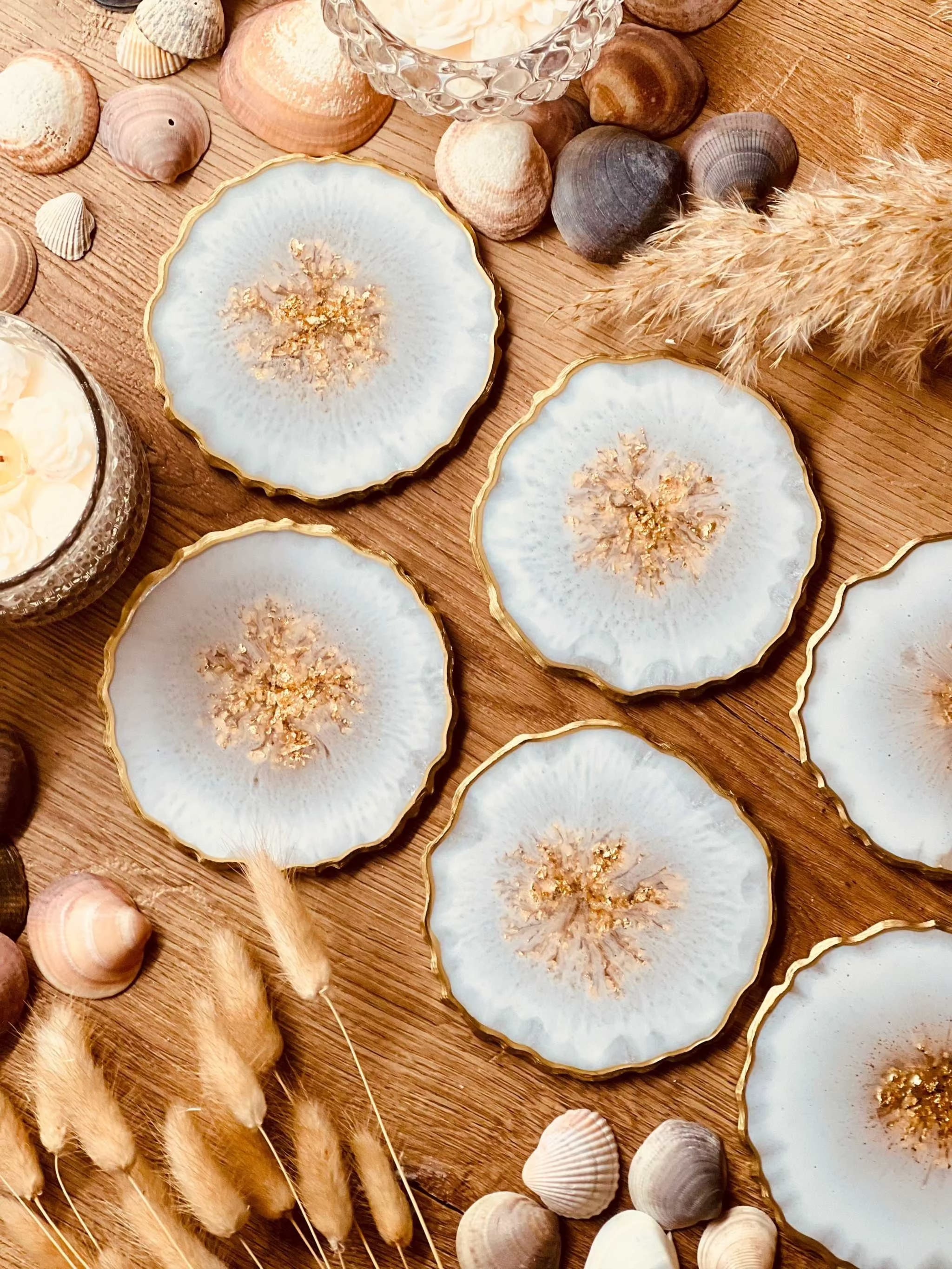 4 elegante handgemachte weiße Resin Untersetzer mit goldenen Details auf Holztisch mit Muscheln und Deko